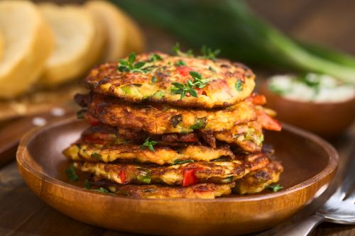 Vegetable and egg fritter made of zucchini, red bell pepper, eggs, green onions and thyme piled on a wooden plate with baguette slices and yogurt dip in the back (Selective Focus, Focus on the front of the thyme sprig on the top of the fritters and on the front of the top fritters)
