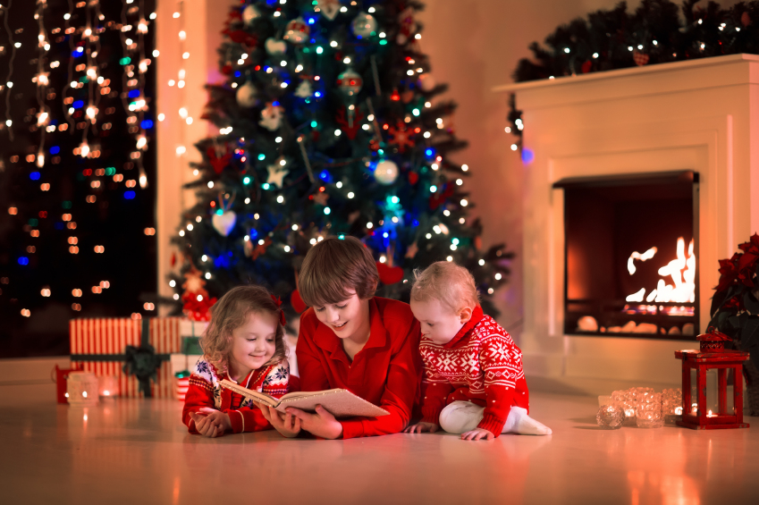 Children read a book and open gifts at fireplace on Christmas eve. Family with child celebrating Xmas. Decorated living room with tree, fire place, candles. Winter evening at home for parents and kids